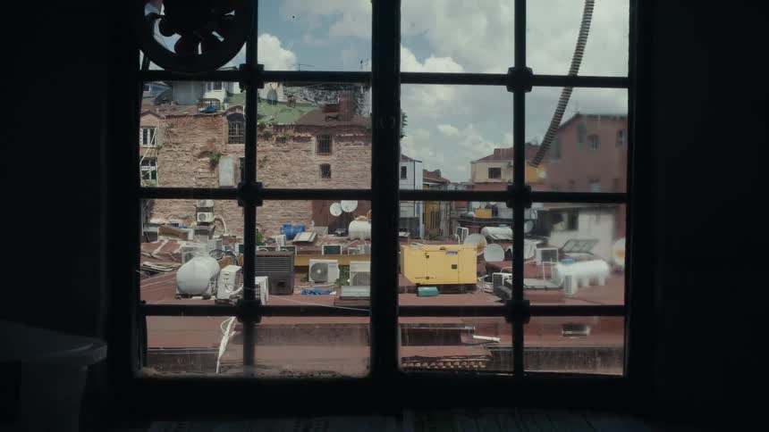 Indoor view looking through a multipaned window at a crowded rooftop in a cityon the roof various objects are visible including satellite dishes air conditioning units and a prominent yellow generator or equipment box