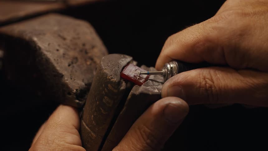 A close up shot of a jewelers hands using a small rotary tool to carefully carve a design into a square red gemstone held securely in a wooden clamp