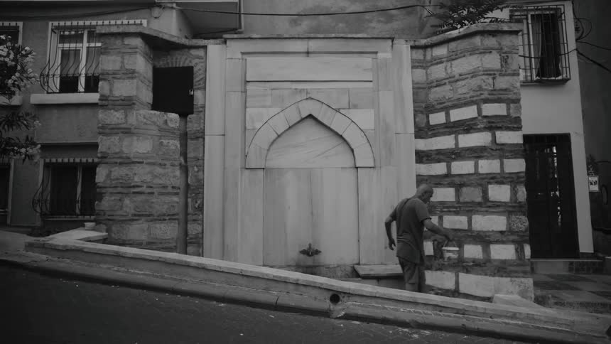 A black and white view shows a man filling a white bucket with water from an old stone fountain built into a brick wall along a sloped city street