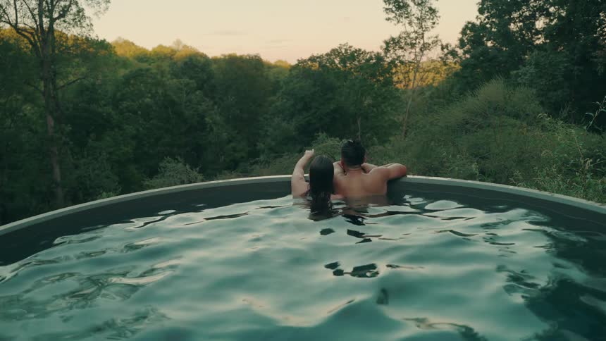 Couple relaxes in elevated pool surrounded by forest at sunset