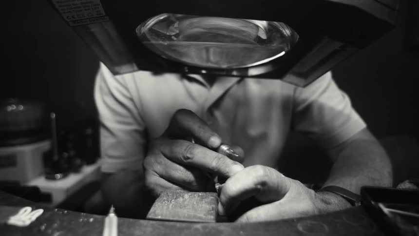 Craftsperson working on a metal piece with a precision tool at a workbench using a magnifying visor