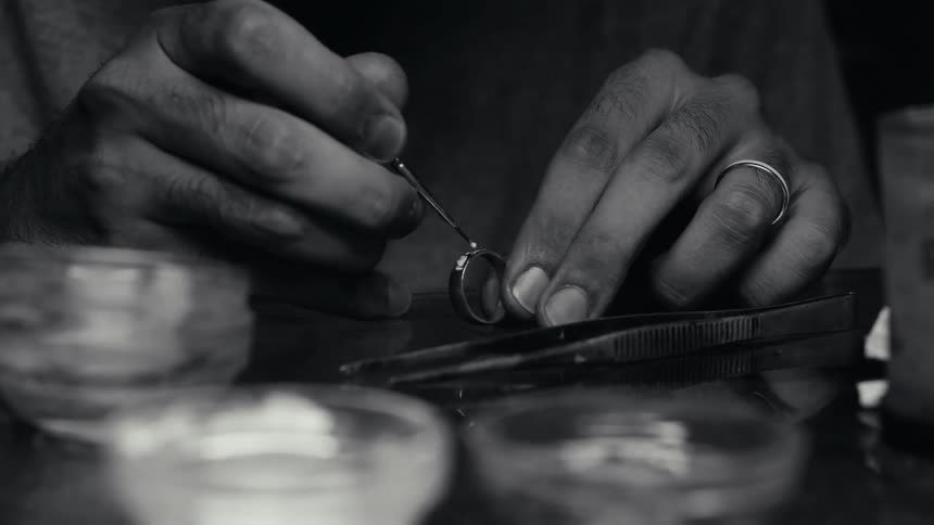 A jeweler carefully uses a fine tool to apply a substance or adjust a setting on a ring held between their fingersthe black and white footage highlights the intricate detail of the workspace including small containers and tweezers laid out on the ben