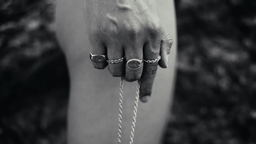A black and white view shows a persons hand wearing several rings and holding a long chain against a dark background