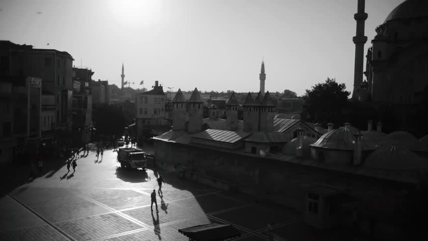 A black and white view from above shows several people walking across a large open plaza in a city with several buildings and a mosque in the background under a bright sun