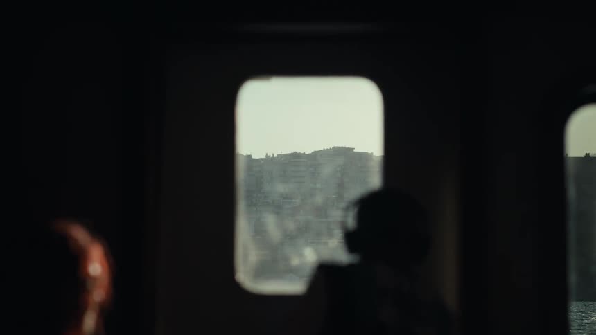 A blurry silhouette of a person looks out of a small boat window toward a distant city the camera moves slowly through the dark interior of the vessel as more figures appear in shadow