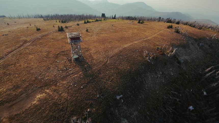An aerial view shows a person riding a motorcycle across a dry grassy mountain ridge past a tall wooden lookout tower
