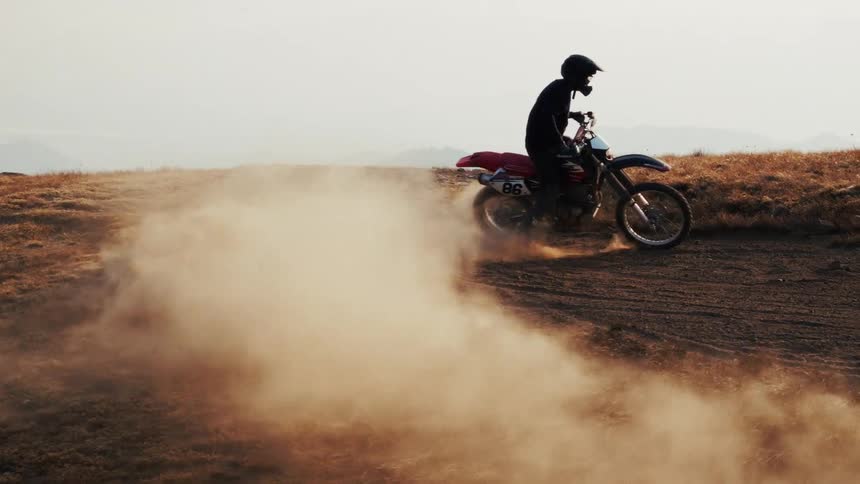 Motorcyclist speeds along dry trail with low vegetation and dust cloud