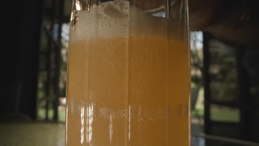 Orange liquid with foam accumulation being poured into a transparent glass container in a lit environment