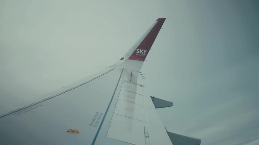 Looking out from a cabin window the camera captures the white wing of a plane as it cuts through a dense layer of gray clouds eventually revealing a vast flat landscape and a hazy horizon during a steady flight