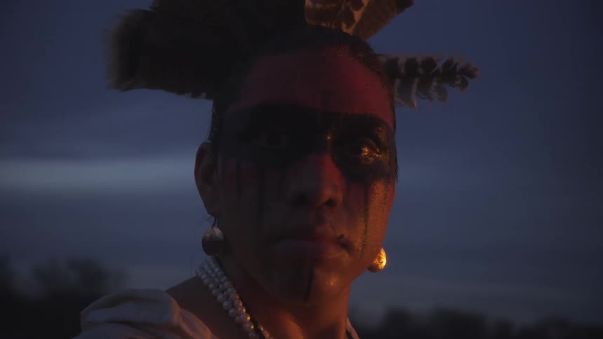 Face of a young adult man wearing native face paint and jewelry with feathers on his head with trees on the background during dusk or dawn