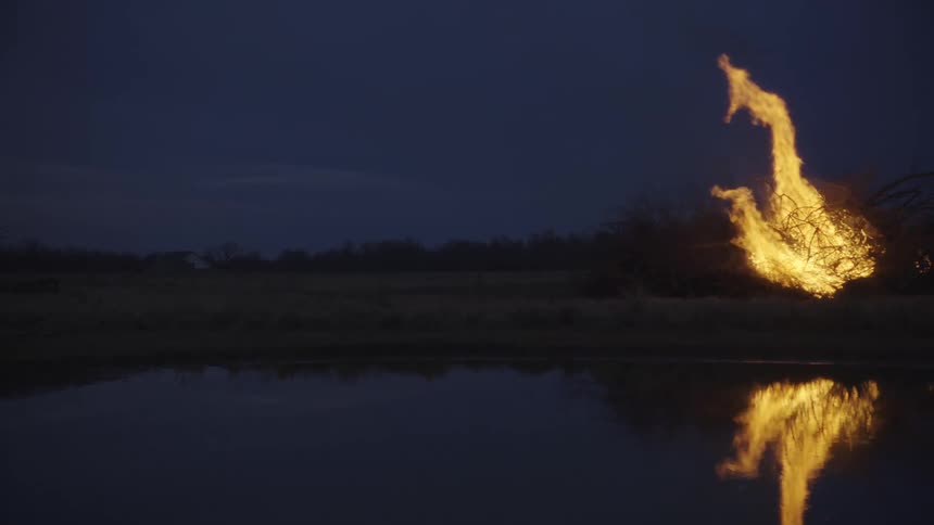 A large bonfire burns intensely in a field at night casting a bright orange glow and a clear reflection onto the surface of a still pond in the foreground while a dark overcast sky hangs over the quiet rural landscape