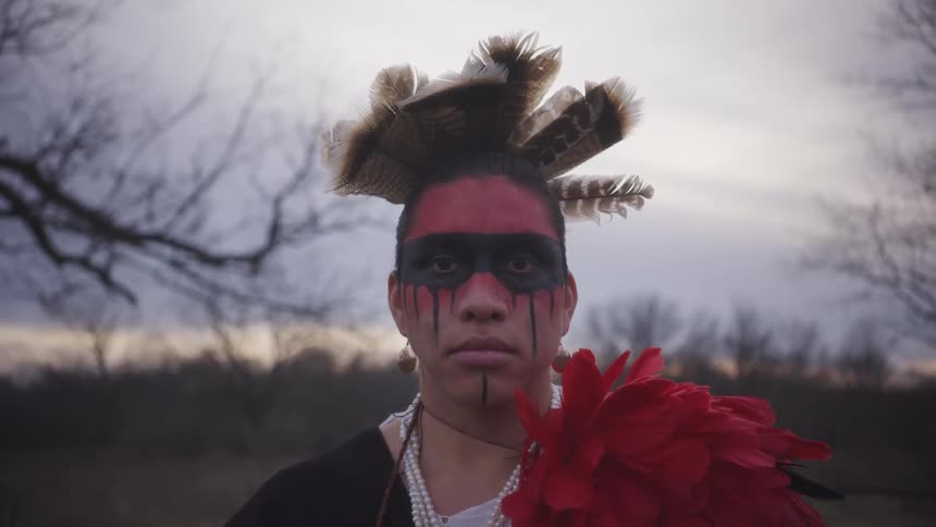 A medium shot shows an indigenous person looking directly at the camera while wearing a feathered headdress necklaces a large red flower and red and black face paint