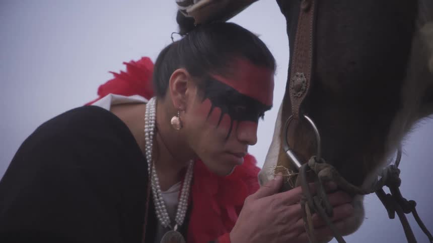 Man wearing feathers on his head and tribal makeup with his hand on his horses muzzle