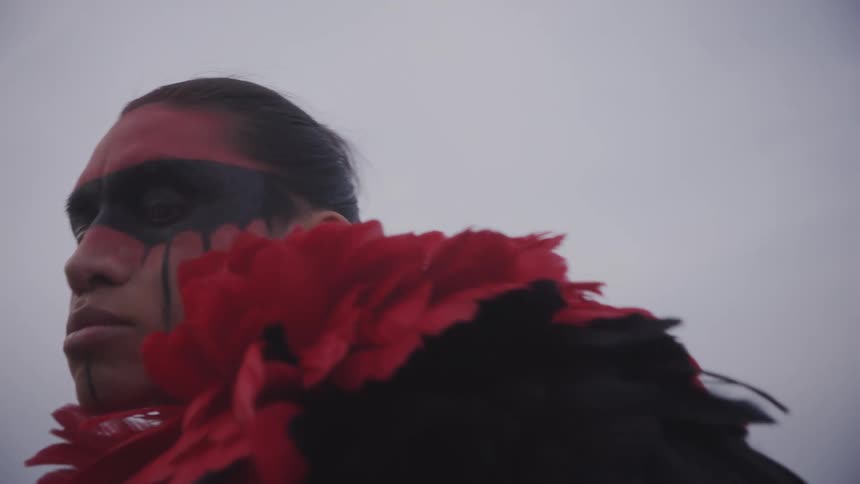 A person grooms a native american with striking red and black face paint under a cloudy sky