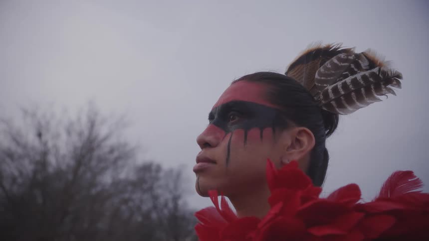 An indigenous man with red and black face paint and a feathered headpiece looks toward the sky