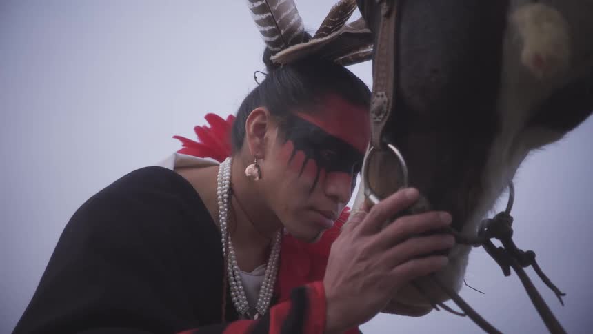 A person in traditional indigenous attire leans their forehead against a horses head in a moment of quiet connection they gently cradle the horses muzzle with their hands while feathers and pearls adorn their hair and neck