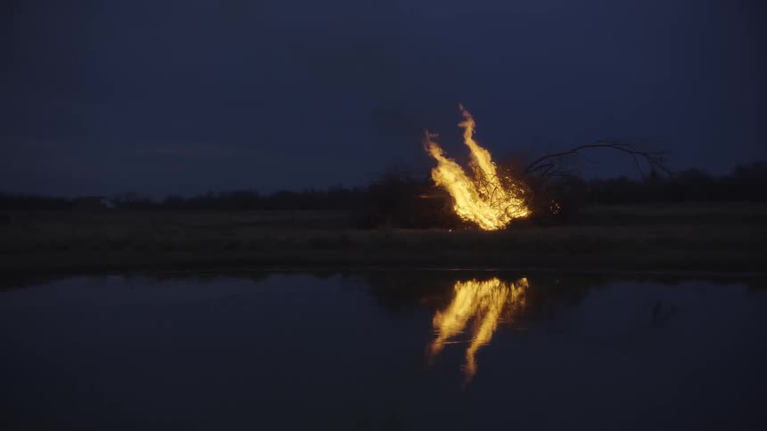 A large brush fire burns brightly in an open field at night casting a vivid orange reflection onto the calm surface of a pond in the foreground while a dark overcast sky hangs over the rural landscape