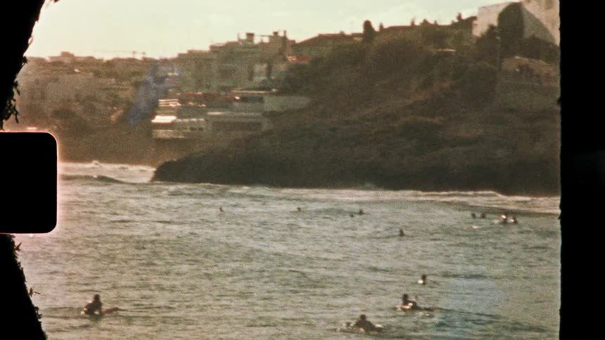 Buildings on mountains overlooking the coast as people swim in the water as waves roll in to shore