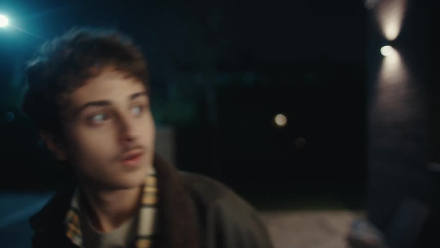 A man in a plaid shirt stands outside at night under a bright light about to kiss before turning and walking away