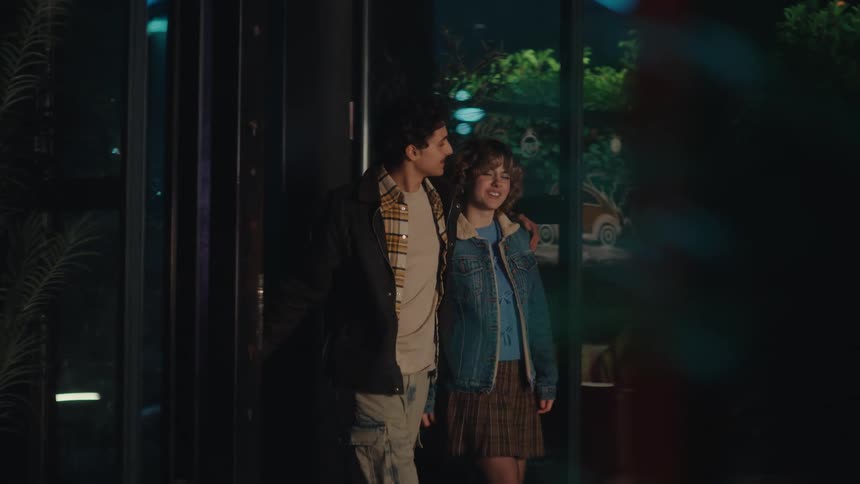 Couple walking side by side at night with an arm over the shoulders in front of an illuminated glass facade