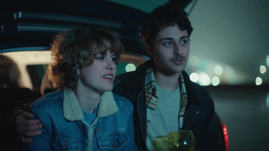 Young couple sitting together on the tailgate of a car along the coast talking and holding a glass of champagne