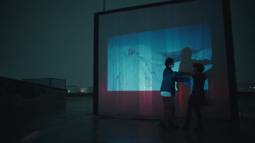 Couple dancing in a nighttime outdoor area in front of a luminous projection on a rectangular structure