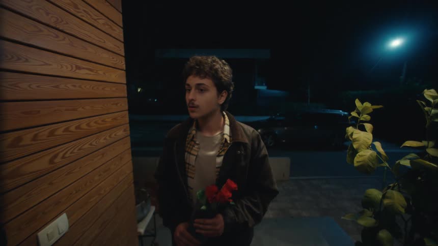 A young man stands at a front door holding a bouquet of red roses at night