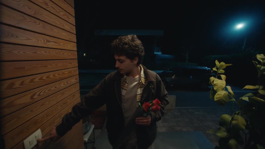 A man holding a bouquet of roses rings a doorbell and waits outside a house at night