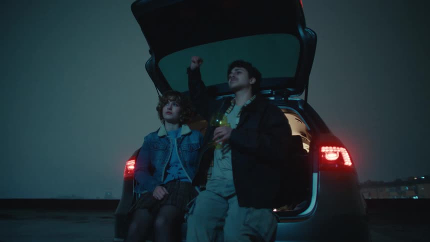Couple sitting in the open trunk of a car at night holding bottles with rear lights on
