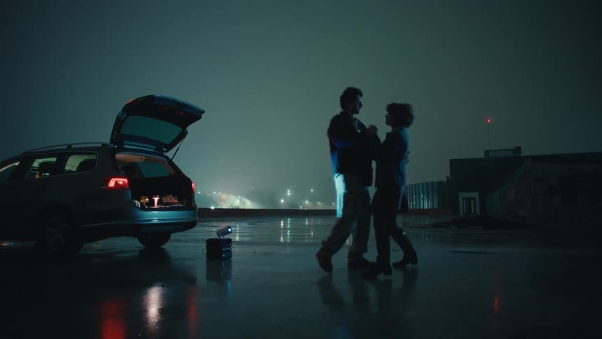 A man and a woman dance on a wet rooftop at night next to a car with its trunk open