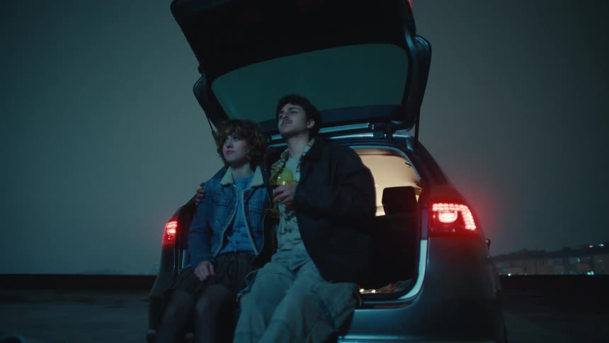 A couple enjoys a romantic toast on the open car trunk overlooking a scenic night view