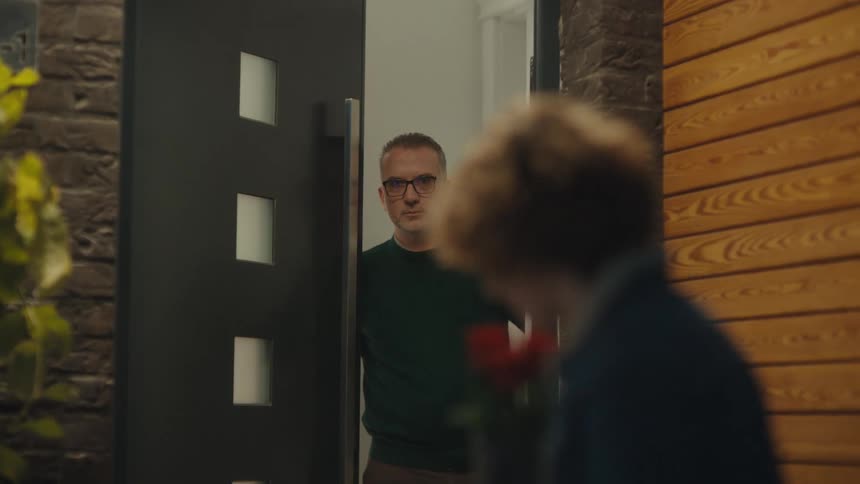 Couple is standing and conversing indoors at night with the woman holding flowers being invited inside