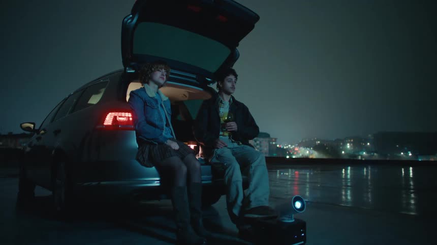 Man and woman cuddle sitting in the trunk of a car while drinking wine with a city behind them in the distance