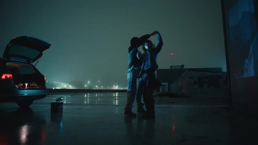 Two people dancing in a wet outdoor area at night with an open car trunk and city lights in the background