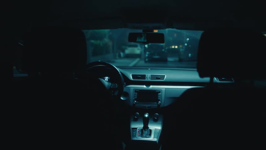 A man and a woman sit in the front seats of a car at night before the woman exits the vehicle and walks toward a building