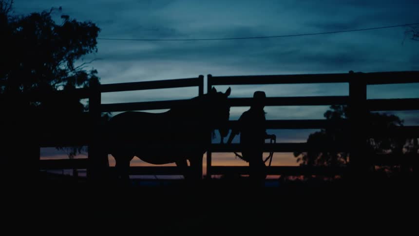 Silhouetted against a deep blue twilight sky a rancher calmly leads a horse along a wooden fence as the last strip of sunset glows on the horizon