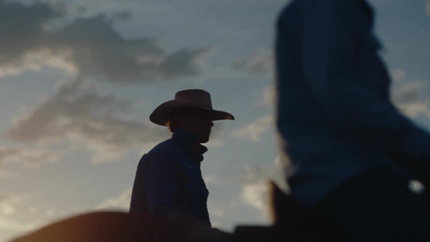 Silhouetted cowboys ride horseback against a vibrant sunset sky