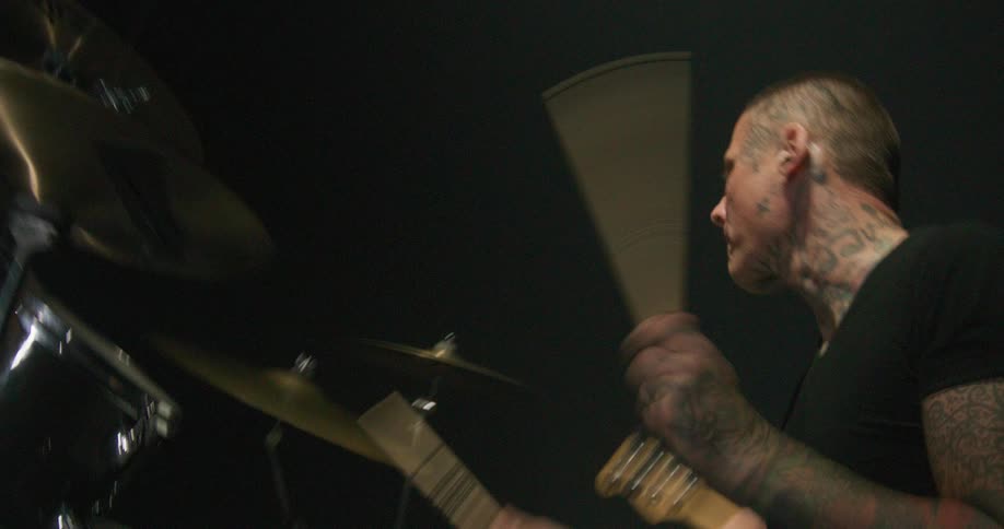Adult man wearing dark clothes playing the drums next to another adult man playing an electric guitar on a stage at a music concert