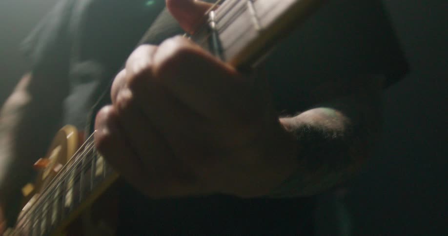 A musicians hand firmly grips the neck of a goldtop electric guitar fingers sliding along the fretboard while the other hand strums in a dark atmospheric setting