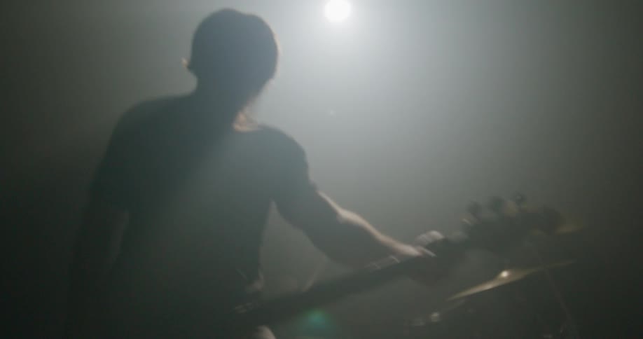 A male musician in silhouette performs on a dark stage filled with hazy smoke and bright backlighting as he aggressively plays an electric guitar while standing in front of a drummer during a live concert set
