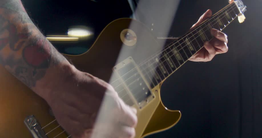 A man with tattooed arms plays a gold top electric guitar in a dark studio