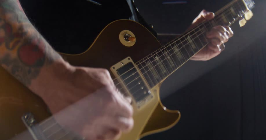 A close view shows a musician with tattooed arms playing a gold electric guitar in a dark setting