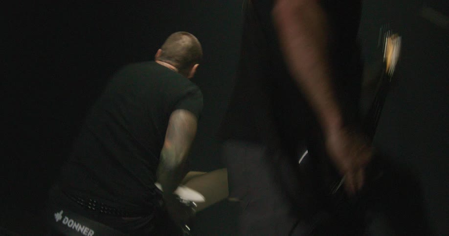 A drummer with tattoos on his arms plays a drum set while a bass player performs beside him in a dark room