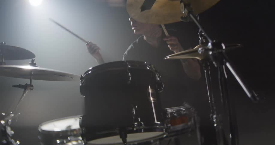 Drummer playing in a dark studio seen in closeup between cymbals and drums under directional lighting