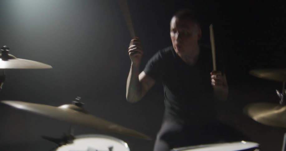 Musician playing guitar in a dark studio with directional lighting and a partially visible drum kit nearby