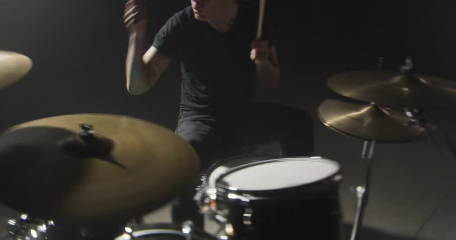 A man with tattoos plays a drum set energetically in a dark room under a single bright light