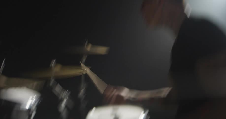 A tattooed man in a dark hazy studio vigorously plays a drum kit striking the cymbals and snare with sticks while the camera moves around him to capture the intensity of his performance under a bright spotlight