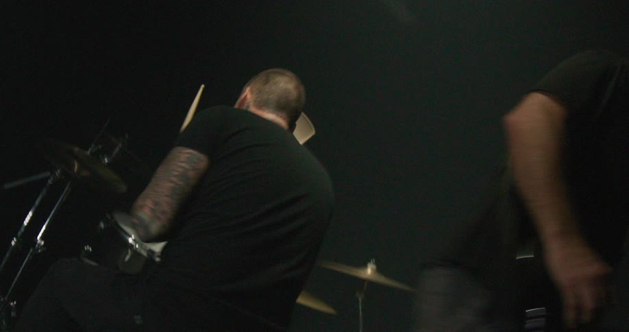 A heavily tattooed drummer intensely plays a black drum kit under dim moody stage lightingthe camera then pans across to show a bassist in a black tshirt energetically performing alongside him
