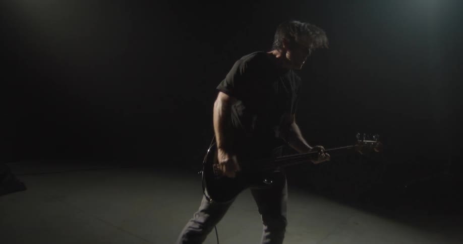 A bassist in a black shirt performs with high energy under a dramatic spotlight in a dark studio he jumps and headbangs while playing his instrument creating a dynamic and moody atmosphere