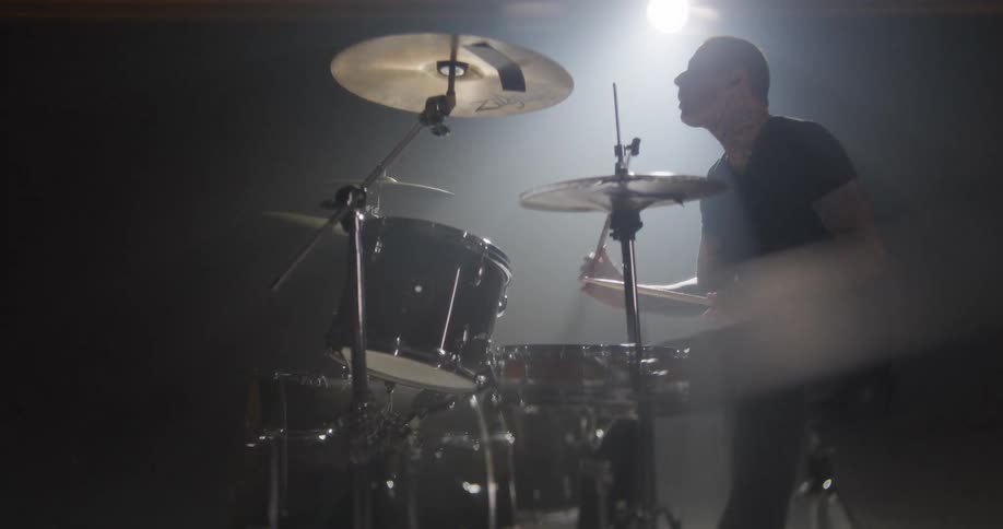 A man with tattoos plays a drum set energetically in a dark room under a single bright light
